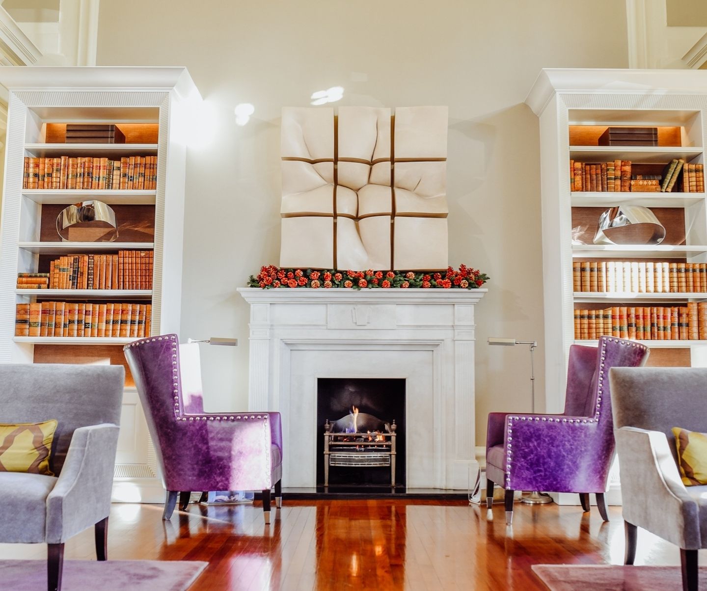 A lounge with two purple chairs facing each other in front of a fireplace with shiny wooden floor and bookcases either side of the fireplace